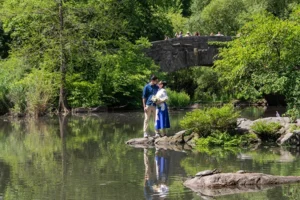 The couple by a stone bridge in Central Park, greenery and pond behind them