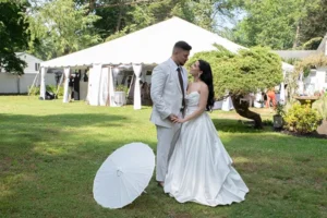 Bride in a satin ball gown and groom in a light suit on the lawn beside a white parasol, reception tent behind them.