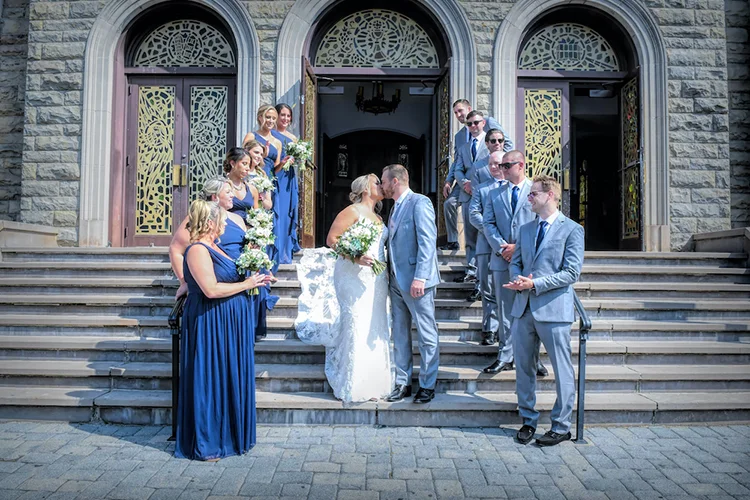 Wedding party on the church steps as the couple kisses, before the Forest Lodge reception