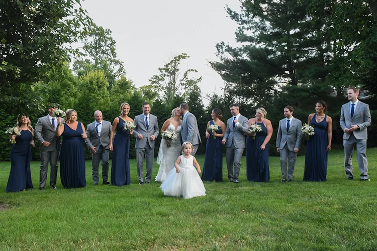 Full wedding party on the lawn as the bride and groom kiss at Forest Lodge
