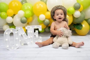 One year old beside a lit ONE marquee sign with a yellow and green balloon garland